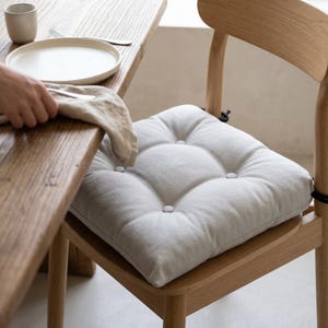 Puede incluir: Un cojín de asiento cuadrado de color gris claro con cuatro botones forrados de tela, sujeto a una silla de madera con lazos negros. El cojín presenta una textura mate suave y una construcción acolchada diseñada para la comodidad al sentarse.