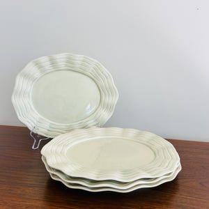 May include: A stack of four off-white ceramic oval plates with a wavy, textured rim. One plate is displayed on a clear stand. The plates are on a dark wood surface against a white background.