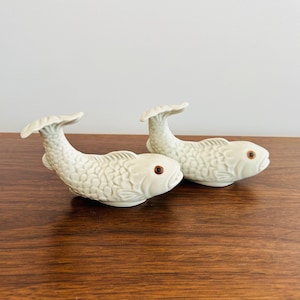 May include: Two ceramic fish figurines with a textured scale pattern and upturned tails. The fish are a pale green color with brown eyes. The figurines are displayed on a wooden surface.