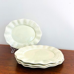 May include: A set of four round ceramic plates in a pale, glossy sage green finish. Each plate features a distinct wavy, scalloped edge design that creates a rippled texture around the rim, while the center of each plate remains smooth and flat.
