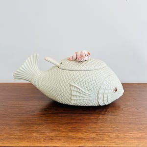 May include: A ceramic fish-shaped serving dish with a lid and a spoon. The dish is light green with a textured scale pattern and a small, pink and white seashell accent on the lid. The fish has a brown eye.