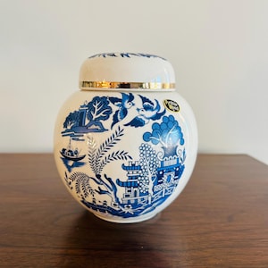 May include: A white and blue ceramic jar with a lid, featuring a classic Willow pattern design. The jar has a gold rim and the text "Arthur Wood" on a sticker. The design includes trees, birds, and a boat.