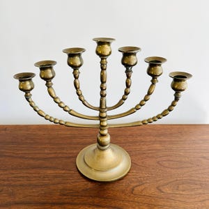 May include: A brass menorah with seven candle holders. The base is round, and the arms curve upwards, each holding a cup-shaped candle holder. The metal has a warm, aged patina, suggesting it is vintage or antique.