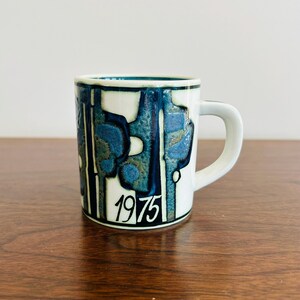 May include: White ceramic mug with a blue and teal abstract design. The mug has a white handle and the year "1975" printed in black. The mug is on a wooden surface.