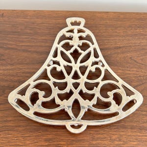 May include: Silver-colored metal trivet in a bell shape with an intricate openwork design. The trivet has a decorative top and a rounded bottom edge. It is placed on a dark brown wooden surface.