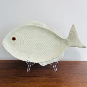 May include: A large, off-white ceramic fish-shaped platter. The platter has a textured scale pattern and a detailed tail fin. The fish has a brown eye and is displayed on a clear acrylic stand. A decorative serving dish.