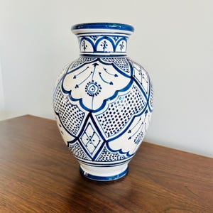 May include: A white and blue ceramic vase with intricate geometric patterns. The vase features a bulbous body, a narrow neck, and a flared rim. The design includes dots, diamonds, and floral motifs, all in a deep blue color. The vase is a decorative item.