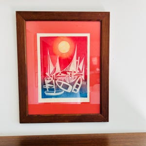 May include: Framed artwork featuring a nautical scene with sailboats and a sun. The artwork has a red and blue background with white sailboats. The frame is made of dark brown wood. The artwork is displayed on a white wall.