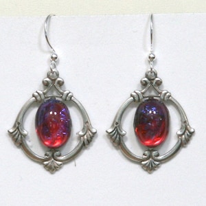 May include: A pair of silver-toned earrings with an oval, red and purple iridescent glass stone set within a decorative frame. The frame has an ornate, openwork design with curved lines and floral accents. The earrings hang from a simple hook.