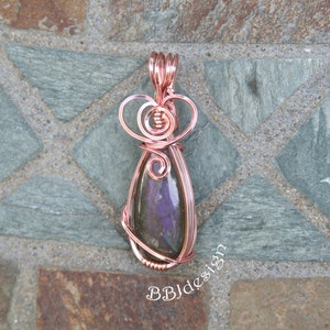 May include: A copper wire pendant with a purple gemstone in a teardrop shape. The wire is carefully wrapped around the stone, with decorative swirls at the top. The pendant is displayed on a textured, stone-like surface. The text "BBJdesign" is visible.
