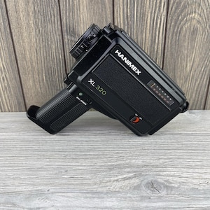 May include: Black Hanimex XL 320 vintage movie camera. The camera has a textured body, a lens, and a handle. The text "HANIMEX" and "XL 320" are visible on the camera body. The camera has a red on/off switch and a battery check light.