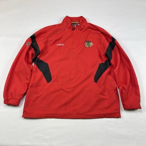 May include: Red and black Chicago Blackhawks Reebok jacket. The jacket has a half-zip closure, long sleeves, and the team logo on the chest. Black panels are on the sides. The Reebok logo is on the chest.