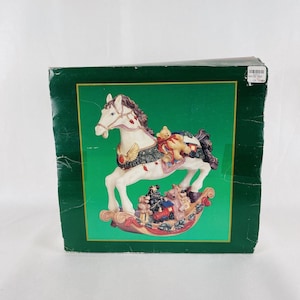 May include: A decorative rocking horse figurine with a white body, brown mane, and a red and gold saddle. The horse is adorned with a teddy bear and other small toys. The figurine is displayed on a green box.