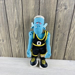 May include: A blue plush toy alien wearing a black and yellow basketball uniform. The alien has large ears, a grumpy expression, and white hair. The uniform has the number "0" on the front.
