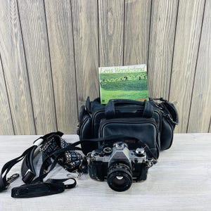May include: A black Canon AE-1 camera with a lens, a black camera bag, and a book titled "Lens Wonderland" are displayed. A patterned camera strap is also present. The items are on a light gray surface.