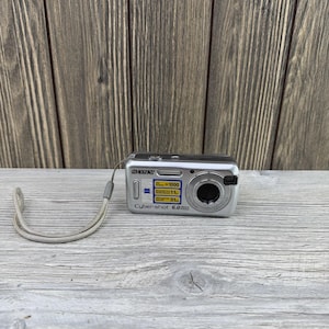 May include: A silver Sony Cyber-shot 6.0 megapixel digital camera with a Zeiss lens, a built-in flash, and a gray wrist strap. The front of the camera features a blue and yellow sticker displaying technical specifications and the Sony brand name.