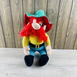 Può includere: Un peluche di Yosemite Sam, un personaggio dei cartoni animati. Il giocattolo ha una barba e un cappello rossi, una camicia gialla, pantaloni blu e stivali neri. Il cappello è verde con un bordo rosso. La cintura è nera con una fibbia dorata.