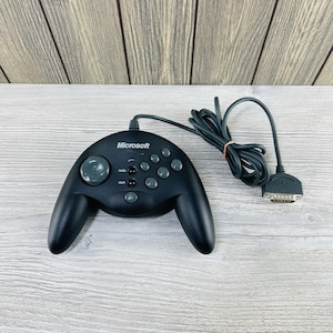 Puede incluir: Un mando Microsoft Sidewinder negro con una cruceta gris y botones grises. El mando tiene un cable negro largo con un conector. La palabra "Microsoft" está impresa en la parte superior del mando.