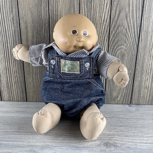 May include: A Cabbage Patch Kids doll with a bald head, brown eyes, and a smiling expression. The doll is wearing a black and white checkered shirt under blue denim overalls. The overalls have a green and white patch.