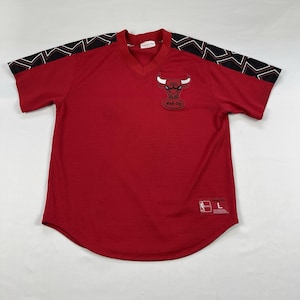 May include: Red Chicago Bulls jersey with a V-neck and short sleeves. The jersey features the Bulls logo on the chest and black and white geometric patterns on the shoulders. The jersey has a size L tag.