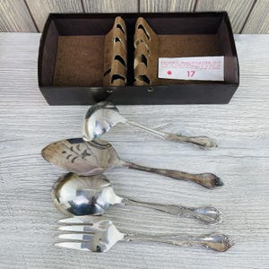 May include: Silverware set including a ladle, serving spoon, soup spoon, and fork. The utensils have ornate handles and are arranged on a light wood surface. A black box with brown dividers is in the background.