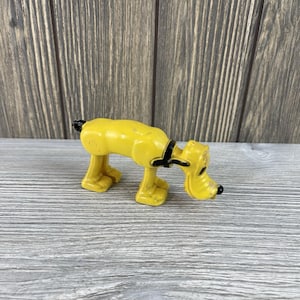 Könnte beinhalten: Eine gelbe Pluto-Figur mit schwarzen Akzenten, darunter Halsband und Ohr. Der Hund befindet sich in einer Schnüffelpose, mit der Nase nahe am Boden. Das Spielzeug ist aus Kunststoff und hat ein Vintage-Aussehen.