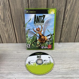 May include: An Xbox video game case and disc for Antz Extreme Racing. The case features a winged ant character against a green grass background with the title text. The circular disc is white and green with the game title and Xbox logo printed.