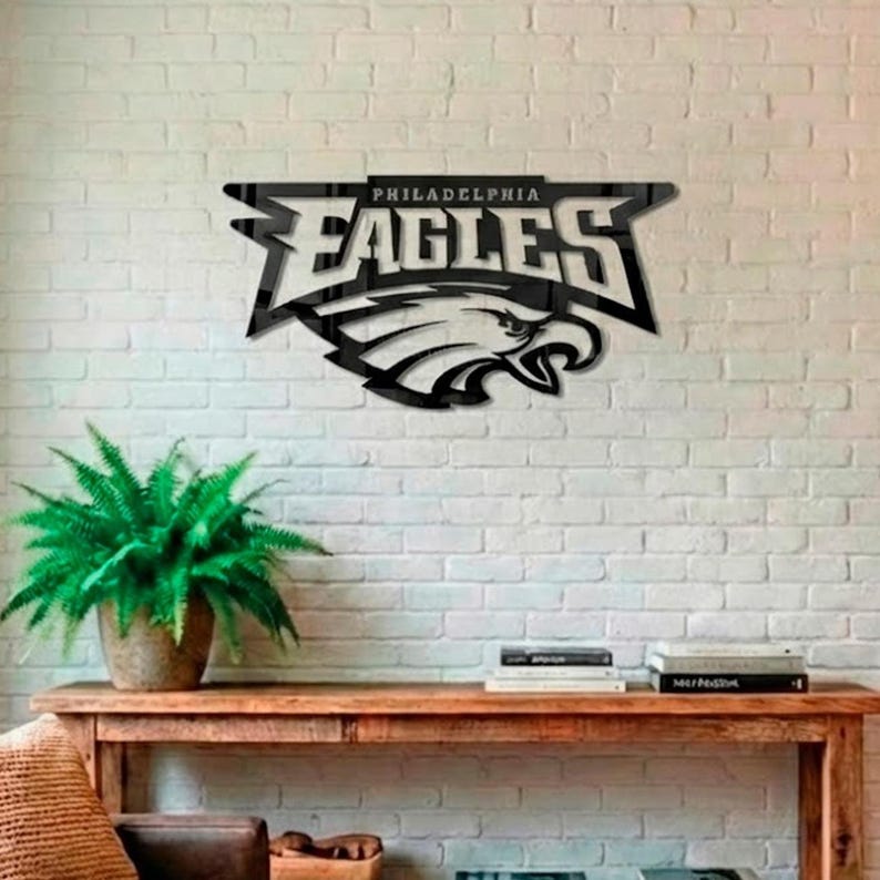 Puede incluir: Arte mural de metal negro con el logotipo de los Philadelphia Eagles. El dise&ntilde;o incluye el nombre del equipo y una cabeza de &aacute;guila detallada. La obra de arte est&aacute; montada en una pared de ladrillo blanco.