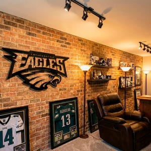 Puede incluir: Una habitaci&oacute;n con una pared de ladrillo decorada con recuerdos de los Philadelphia Eagles. Incluye un gran logotipo de los Eagles de metal negro, camisetas enmarcadas y una camiseta enmarcada con el nombre de Agholor y el n&uacute;mero 13. Un sill&oacute;n reclinable de cuero marr&oacute;n est&aacute; en la esquina.