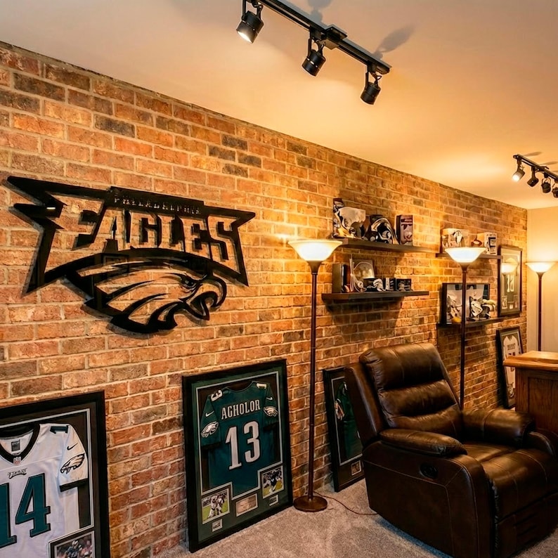 Puede incluir: Una habitaci&oacute;n con una pared de ladrillo decorada con recuerdos de los Philadelphia Eagles. Incluye un gran logotipo de los Eagles de metal negro, camisetas enmarcadas y una camiseta enmarcada con el nombre de Agholor y el n&uacute;mero 13. Un sill&oacute;n reclinable de cuero marr&oacute;n est&aacute; en la esquina.