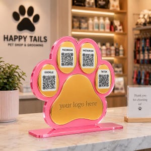 May include: A pink and gold paw-shaped acrylic sign featuring four QR codes labeled Facebook, Instagram, Google, and TikTok. The center gold section includes the text your logo here. The sign stands on a rectangular base.