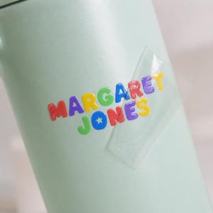 May include: A light green tumbler featuring a clear adhesive strip over a colorful name decal. The text reads MARGARET JONES in multi-colored, bubble-style letters, with a small white star detail inside the letter O in the name JONES.