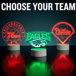 May include: Three illuminated team logo night lights. The logos are for the Philadelphia 76ers, Eagles, and Phillies. The text "CHOOSE YOUR TEAM" is at the top. The lights are red and green.