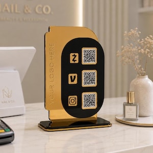 May include: A gold and black acrylic QR code display stand. The stand features three QR codes, social media icons, and the text "YOUR LOGO HERE". A perfume bottle and a vase with dried flowers are in the background.