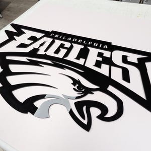 Puede incluir: Un logotipo en blanco y negro de los Philadelphia Eagles, con el nombre del equipo y una cabeza de &aacute;guila estilizada. La palabra "PHILADELPHIA" est&aacute; encima de la palabra "EAGLES". La cabeza del &aacute;guila tiene acentos grises y blancos.