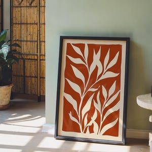 May include: A rectangular framed wall art piece featuring a bold terracotta orange background with a white botanical leaf pattern. The print is enclosed in a thin black frame and displays a series of elongated, organic leaf shapes arranged vertically.