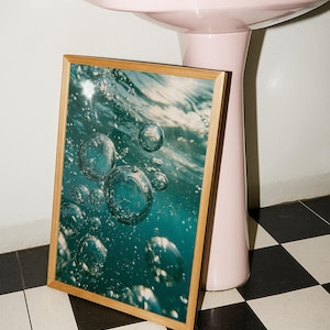 May include: A rectangular wall art print in a light wood frame featuring a high-resolution photograph of underwater air bubbles rising toward the surface. The image displays deep teal and blue water tones with bright light reflections on the bubbles.