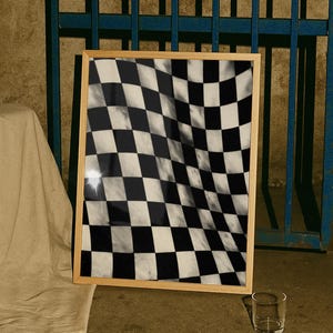 May include: A rectangular wall art piece in a light-toned wooden frame featuring a black and white checkered pattern with a distorted, wavy effect. The print displays a high-contrast grid of squares that appear to ripple across the surface.