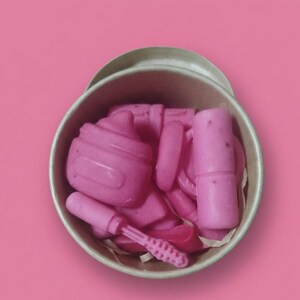 Pode incluir: Uma coleção de sabonetes rosa feitos à mão em forma de itens de maquiagem, incluindo batom, um pincel de maquiagem e um cupcake, dentro de um recipiente redondo marrom claro. O recipiente está em um fundo rosa.