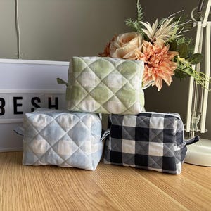 May include: Three quilted fabric cosmetic bags. One is light green with a diamond pattern, one is light blue with a diamond pattern, and one is black and white checkered. The word "BESH" is in a white light box behind the bags.