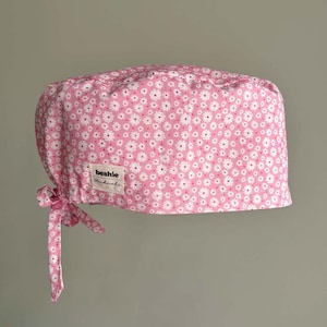 May include: A pink scrub cap featuring a repeating pattern of small white daisies with yellow centres. The cap includes a fabric tie on the side for adjustment and a rectangular white fabric label stitched to the side that reads beshie Handmade.
