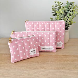 May include: Three pink quilted zipper pouches with white polka dots. The pouches have gold-coloured zips and small white labels reading "beshie handmade". The pouches are in different sizes and are arranged on a wooden surface.