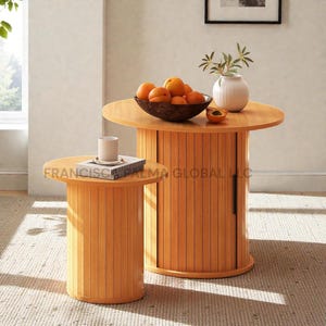 May include: Two wooden side tables with a light brown finish. The tables have a cylindrical base with vertical slats and a round tabletop. One table has a door. The larger table holds a bowl of oranges, a white vase, and a half apricot. The smaller table has a cup and a book.