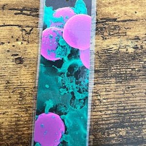 May include: A bookmark with a microscopic image of cells in shades of pink and teal, attached to a gray tassel. The bookmark is rectangular and encased in clear plastic, with a gray string connecting it to the tassel.