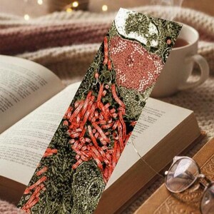 May include: A colorful bookmark with a detailed, abstract design is placed in an open book. The bookmark features red, green, and white patterns. A cup of tea and a pair of glasses rest nearby, creating a cozy reading scene.