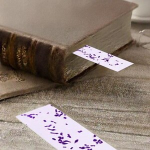 May include: A hardcover book with a brown leather spine and aged pages. A white bookmark with purple abstract shapes is inserted into the book. Another bookmark lies on the wooden surface.