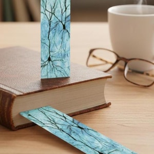 May include: A set of two bookmarks with a neuron design. The bookmarks feature a blue background with black neuron illustrations. One bookmark is standing upright, and the other is lying flat on a book. A cup of coffee and glasses are in the background.