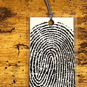 May include: A rectangular bookmark featuring a high-contrast black and white fingerprint pattern. The bookmark is made of a clear material and includes a hole at the top with a silver-colored thread tassel attached to the top edge of the item.