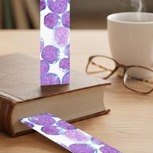 May include: Two bookmarks with a white background and a pattern of purple and pink cell-like shapes. One bookmark is inserted into a brown book, and the other is lying on a wooden surface. A white mug and glasses are in the background.