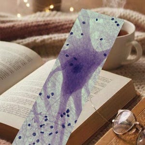 May include: A bookmark featuring a purple neuron illustration is placed in an open book. A cup of tea steams nearby, with a pair of glasses resting on a closed book. The scene is set on a cozy, textured surface, suggesting a relaxing reading environment.